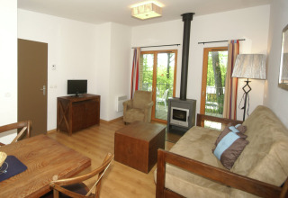 Woonruimte in een lodge met houten meubilair, kachel, tv en grote ramen naar het bosrijke balkon.