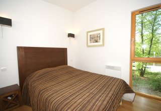 Lichte slaapkamer van een lodge met tweepersoonsbed, houten hoofdsteun en uitzicht op het groene bos buiten.