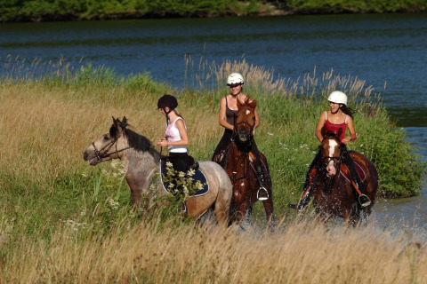 Drie vrouwen maken een paardrijtocht langs een rivier aan een lodge, omringd door hoge grassen.