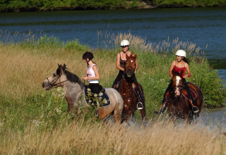 Drie vrouwen maken een paardrijtocht langs een rivier aan een lodge, omringd door hoge grassen.