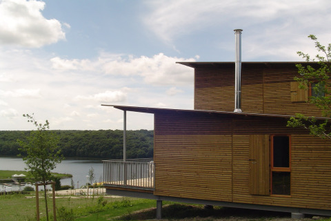Moderne houten lodge met ruime veranda en schoorsteen aan een meer, omgeven door groene natuur.