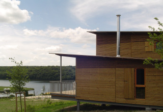 Moderne houten lodge met ruime veranda en schoorsteen aan een meer, omgeven door groene natuur.