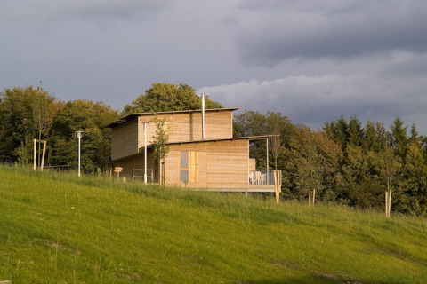 Een eigentijdse houten lodge staat op een grasheuvel met bomen rondom en een bewolkte lucht erboven.
