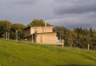 Een eigentijdse houten lodge staat op een grasheuvel met bomen rondom en een bewolkte lucht erboven.