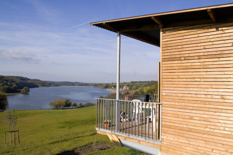 Zicht vanaf een houten lodge met balkon over een rustig meer en glooiende groene heuvels onder een blauwe lucht.