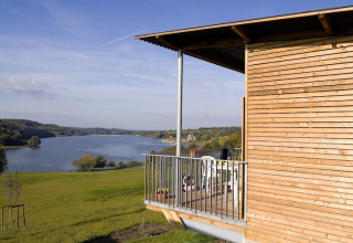 Zicht vanaf een houten lodge met balkon over een rustig meer en glooiende groene heuvels onder een blauwe lucht.