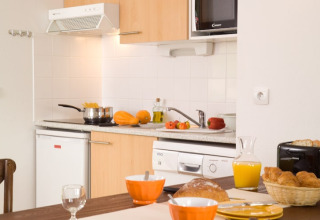 Knusse lodge-keuken met ontbijt op tafel, vers brood, jus d’orange, croissants en groenten in zicht.