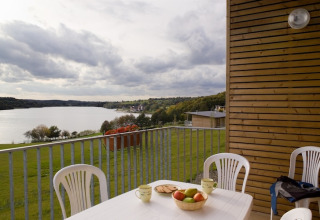 Uitzicht vanaf een lodge op een meer, terras met kunststof stoelen, fruitschaal en kopjes op tafel.