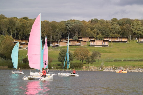 Zeilboten varen op een meer voor houten lodges op een groene heuvel, omringd door bosrijke natuur.