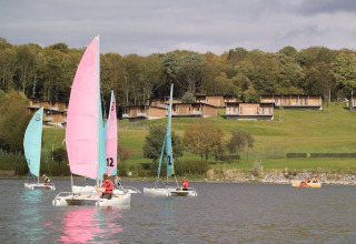 Zeilboten varen op een meer voor houten lodges op een groene heuvel, omringd door bosrijke natuur.