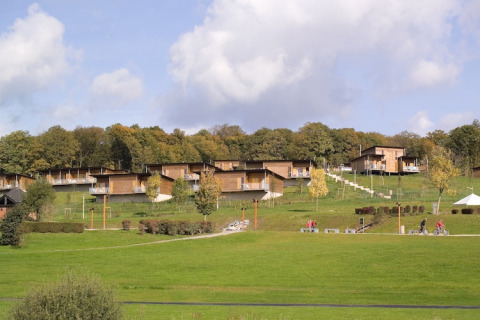 Zicht op moderne houten lodges op een groene heuvel bij bosrand, met fietsers en mensen op de paden onderaan.