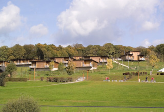 Zicht op moderne houten lodges op een groene heuvel bij bosrand, met fietsers en mensen op de paden onderaan.