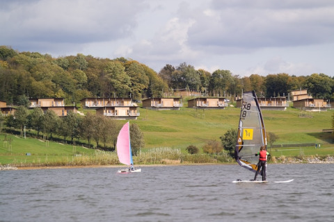 Zicht op lodges aan het meer met mensen die windsurfen en zeilen op een groene heuvel onder de wolken.