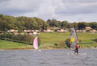 Zicht op lodges aan het meer met mensen die windsurfen en zeilen op een groene heuvel onder de wolken.
