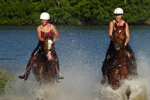 Twee vrouwen met helm rijden op paarden door het water aan een lodge, lachend en genietend van de zon.