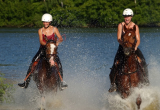 Twee vrouwen met helm rijden op paarden door het water aan een lodge, lachend en genietend van de zon.