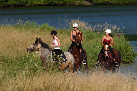 Drie personen te paard rijden langs een meer met hoog gras nabij een lodge tijdens een zomerse dag.