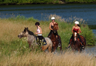 Drie personen te paard rijden langs een meer met hoog gras nabij een lodge tijdens een zomerse dag.