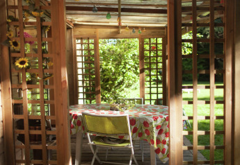 Vista a través de una pérgola de madera a una mesa con mantel floral en el jardín de Maison de vacances - LE PONCHEL.
