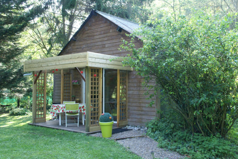 Træhytte med overdækket terrasse og havemøbler, omgivet af grønne planter i Maison de vacances - LE PONCHEL, Frankrig.