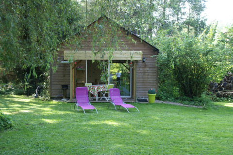 Hytte kaldet Maison de vacances - LE PONCHEL med to lilla liggestole og grøn have omgivet af træer.
