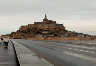 Et foto af Mont Saint-Michel i Frankrig, taget fra vejen, med mennesker gående i forgrunden.