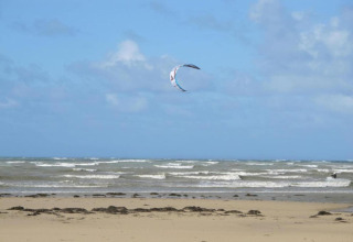 En strand med bølger og en drage i luften ved Maison de vacances - LE PONCHEL, Frankrig, under en blå himmel.