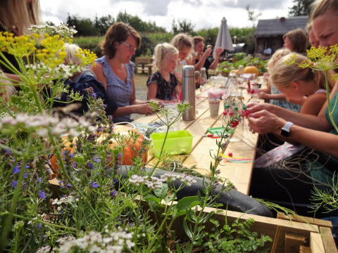 Children and adults enjoy outdoor crafts together at a wooden table surrounded by wildflowers at Campsite Junora.