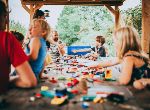 Children and adults playing with toys at a large wooden table under a shelter at Campsite Junora, France.