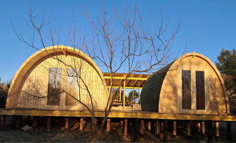 Two arched wooden cabins on stilts at Campsite «Junora» holiday park in Nouvelle-Aquitaine, France at sunset.