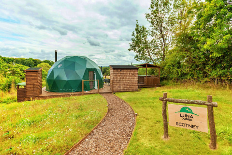 Scotney Luxury Dome, a modern green glamping dome with wooden deck, outdoor seating, and forest backdrop.