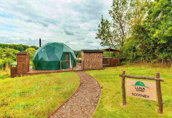 Scotney Luxury Dome, a modern green glamping dome with wooden deck, outdoor seating, and forest backdrop.