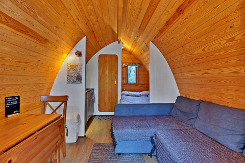 Interieur van een knusse houten pod met bank, eettafel, kleine kitchenette en bed achterin.