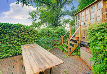 Terrazza in legno con tavolo e casetta presso Higher House, Regno Unito, circondata da vegetazione lussureggiante.