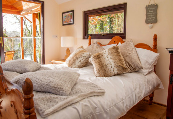 Cozy bedroom inside Yeworthy Eco-Treehouse, UK, featuring wooden bed, fluffy pillows, and balcony view.