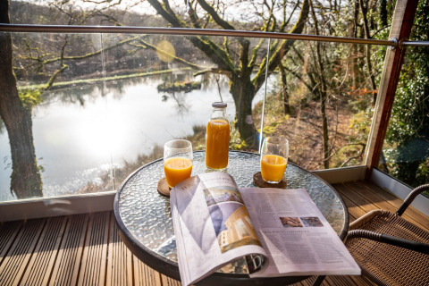 Succo d'arancia e giornale su un tavolo con vista lago a Yeworthy Eco-Treehouse, Regno Unito.