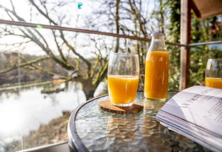 Sole del mattino su tavolo di vetro con succo d’arancia, bottiglia e rivista a Yeworthy Eco-Treehouse, Regno Unito.