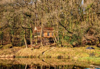 Treehouse by a lake in the woods, Yeworthy Eco-Treehouse UK, with garden furniture and umbrella.