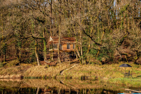 Casa sull’albero vicino a un lago nei boschi, Yeworthy Eco-Treehouse UK, con mobili da giardino e ombrellone.