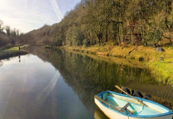 View of a calm lake with a rowboat, forest treehouse, and outdoor seating at Yeworthy Eco-Treehouse, UK.