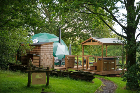 Knole Luxury Dome: Green dome, wooden deck, hot tub, and cozy forest setting with a welcoming entrance sign.