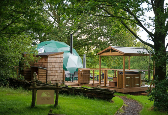 Knole Luxury Dome: Green dome, wooden deck, hot tub, and cozy forest setting with a welcoming entrance sign.