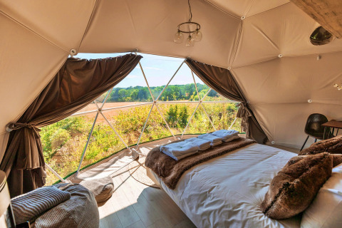 Interior of Scotney Luxury Dome in the UK, showing cozy bed and panoramic window view of countryside.