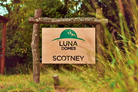 Wooden sign reads 'LUNA DOMES SCOTNEY' with green dome logo in a lush UK outdoor setting.