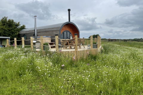 The Campion pod sits on a wooden deck surrounded by wildflowers and fields under a cloudy sky.