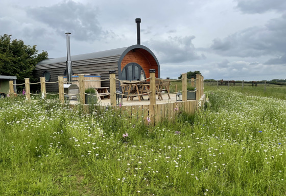 The Campion pod sits on a wooden deck surrounded by wildflowers and fields under a cloudy sky.