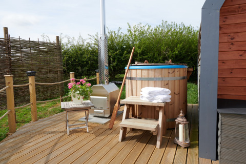 Buitenhouten hottub met trapje, handdoeken, bloemen en lantaarn op terras naast een pod-accommodatie.