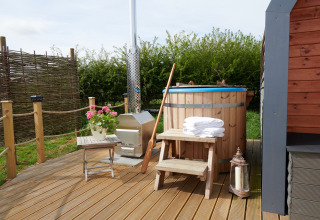 Buitenhouten hottub met trapje, handdoeken, bloemen en lantaarn op terras naast een pod-accommodatie.