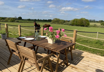 Terrasse en bois avec table, chaises, bouquet fleuri et vue sur des chevaux dans un champ vert au Royaume-Uni.