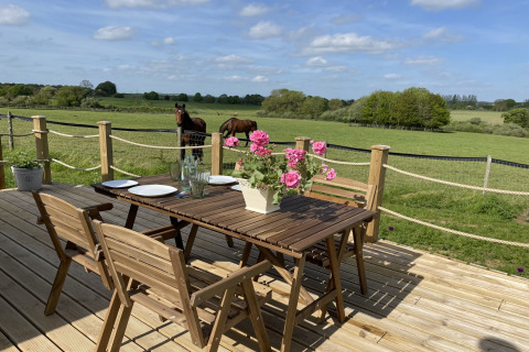 Wooden deck with a table and chairs, fresh flowers, and a scenic view of horses in a green UK field.